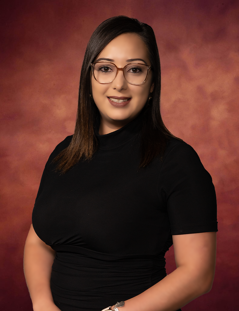 Yennifer Velez, with shoulder-length dark hair and glasses, is smiling at the camera. She wears a black top against a warm-toned, textured background, exuding confidence and charm.
