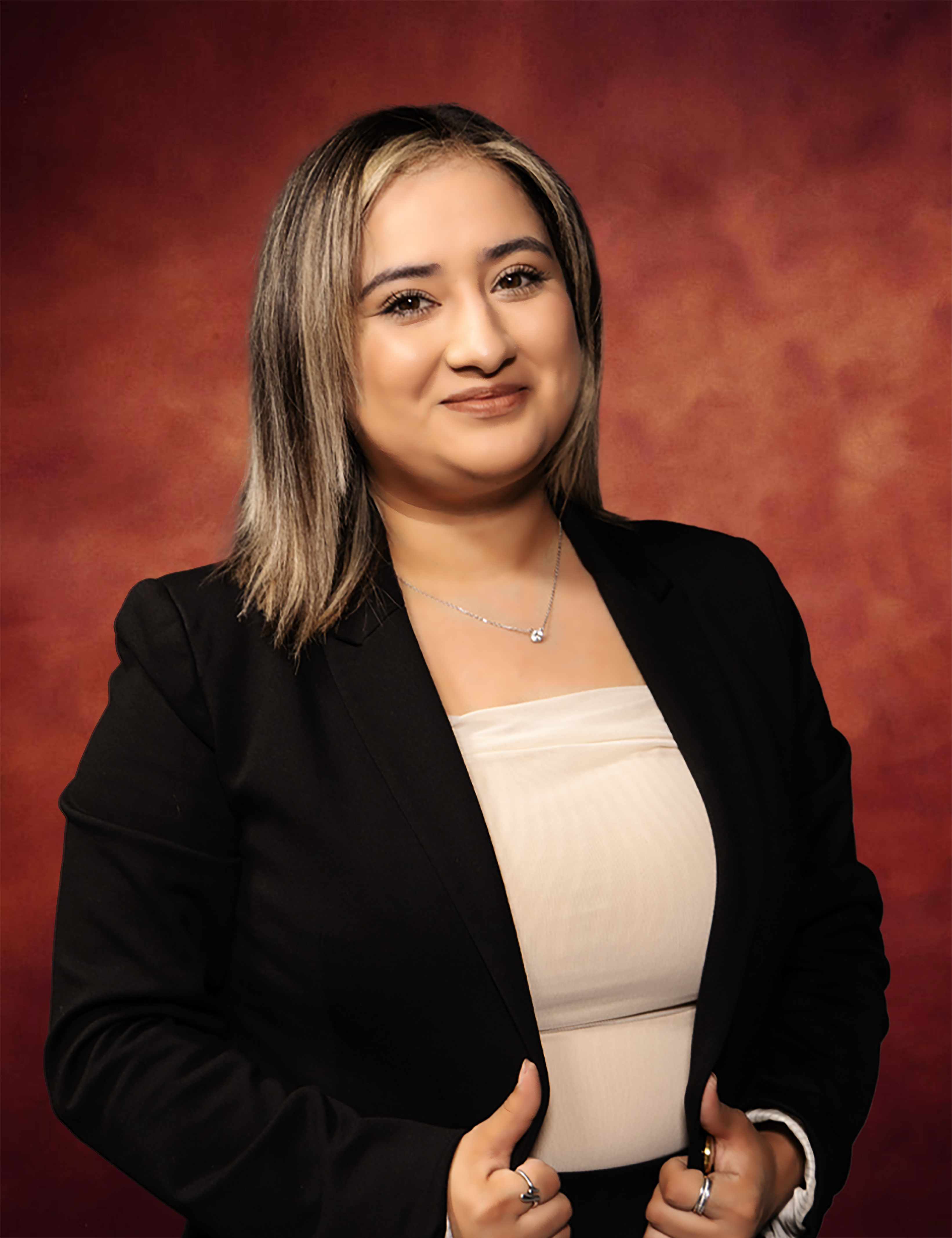 A person with shoulder-length hair and a smile, perhaps reminiscent of Yoselin Delgado, is wearing a black blazer over a light-colored top. They stand against a warm, reddish-brown background.