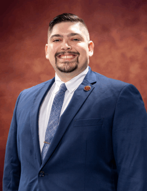 Douglas Alford, a man in a blue suit and tie, stands smiling against a warm, textured brown background. He has short dark hair and a neatly trimmed beard and mustache.