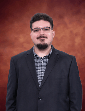 Andrew Vasquez, a man with glasses and a beard, wears a black blazer over a checkered shirt. He stands against a marbled, warm brown background, smiling subtly and looking directly at the camera.