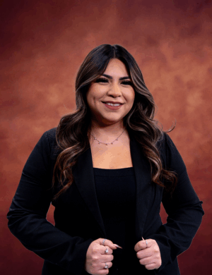 A person with long wavy hair, identified as Abilaisha Marquez, wearing a black jacket stands against a warm brown textured background. They are smiling and have their hands clasped in front.