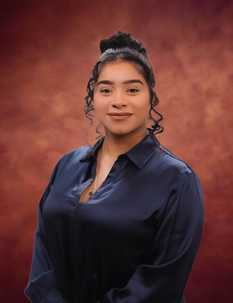 Ashley Ibarra, with dark hair styled in a bun, stands gracefully against a warm, reddish-brown background. She's wearing a dark blue blouse and smiles slightly while facing the camera.