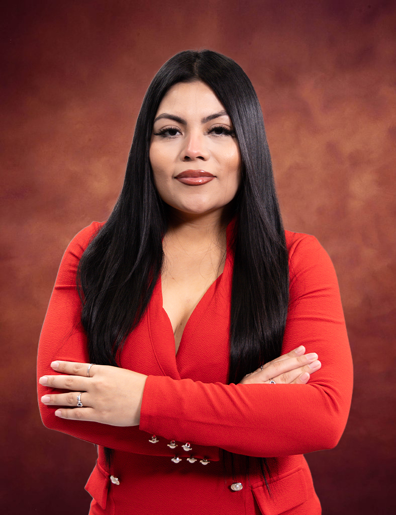Abilaisha Marquez stands confidently, her long black hair cascading over a striking red blazer, arms crossed against a warm brown gradient backdrop.
