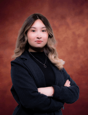 Abilaisha Marquez stands confidently with arms crossed, her long, wavy hair cascading over a dark blazer and a necklace, set against a warm, textured brown background.