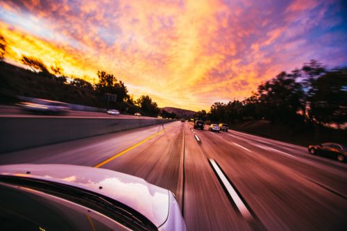A car speeds down a Texas highway at sunset, where the sky is painted with vibrant hues of orange, pink, and purple. Trees line the road, and other vehicles blur past in the distance, highlighting the thrill and consequences of reckless driving.