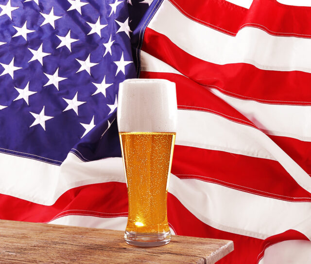 A tall glass of beer with a frothy head sits on a wooden table. In the background, a large American flag with stars and stripes waves proudly, evoking a classic Fourth of July vibe. The light-colored beer appears refreshing, perfectly complementing the patriotic setting.