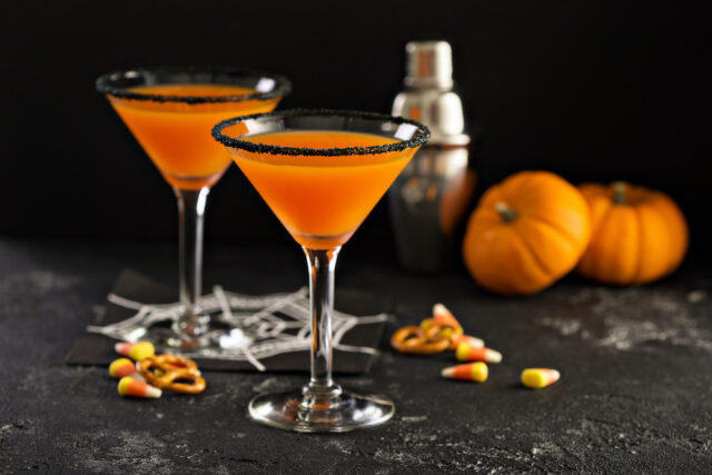 Two orange cocktails with black sugar rims in martini glasses sit on a dark surface, evoking a festive Halloween Night. Nearby are candy corn pieces, pretzels, small pumpkins, and a metal shaker. A white spider web decoration completes the spooky scene.