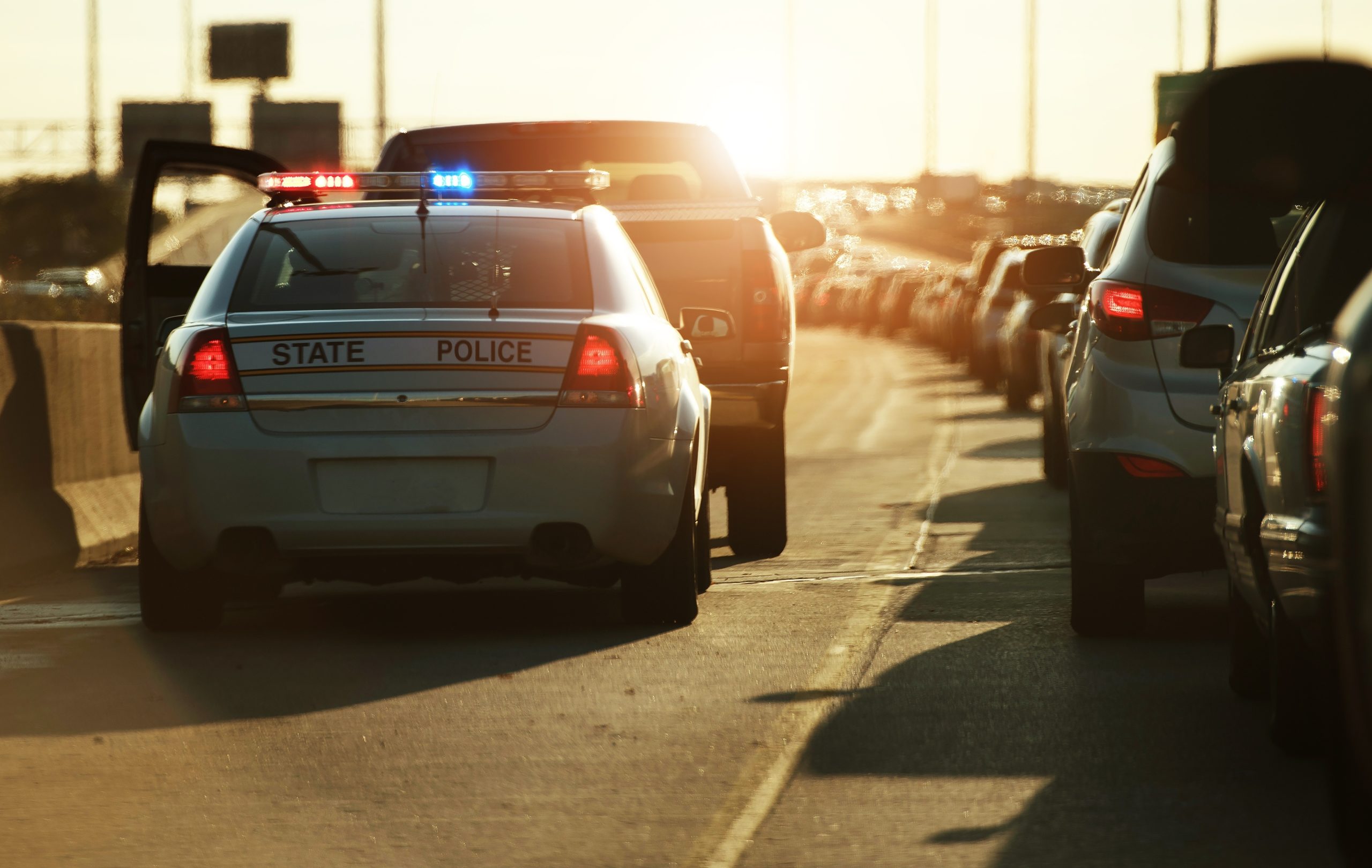 A police car with flashing lights is parked on the shoulder of a busy Texas highway during sunset, addressing a DWI charge. Traffic is backed up in the right lane, with multiple cars visible ahead. The overall scene is backlit by the sun.