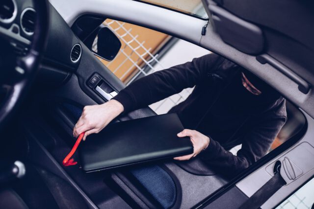 In a shadowy scene typical of theft crimes, a person in dark clothing is reaching into a car in Texas, trying to snatch a black laptop from the passenger seat. The car door stands open, their face partially obscured as they commit this audacious act.