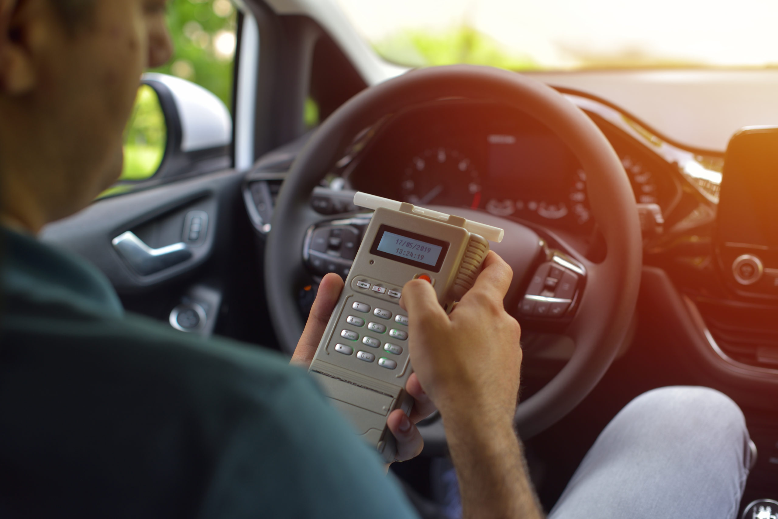 A person is sitting in the driver's seat of a car, holding a vintage mobile phone to their ear. The car's steering wheel and dashboard are visible, with ignition interlock devices in view. Sunlight filters through the window, illuminating the scene.