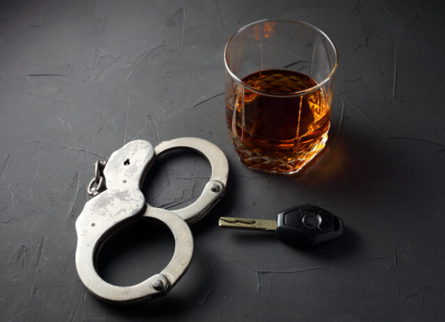 A glass of whiskey, a car key, and a pair of handcuffs sit ominously on a dark textured surface, painting a stark reminder of the consequences of DWI in Texas.