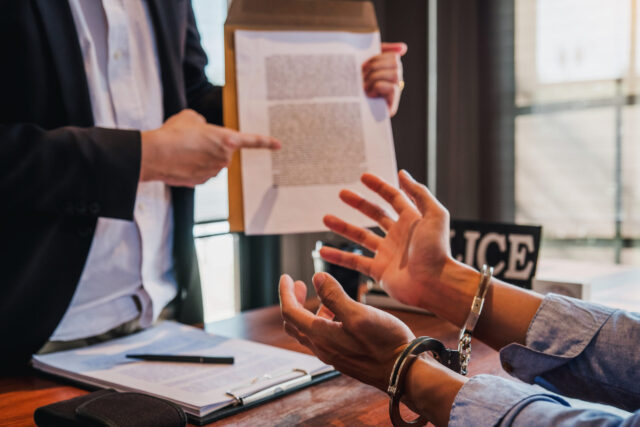 A person in handcuffs is seated at a table with a piece of paper and pen. Nearby, another points at a document on a clipboard, highlighting alternatives to incarceration. The scene unfolds in an office setting, suggesting an interrogation or legal discussion within Texas's justice system.