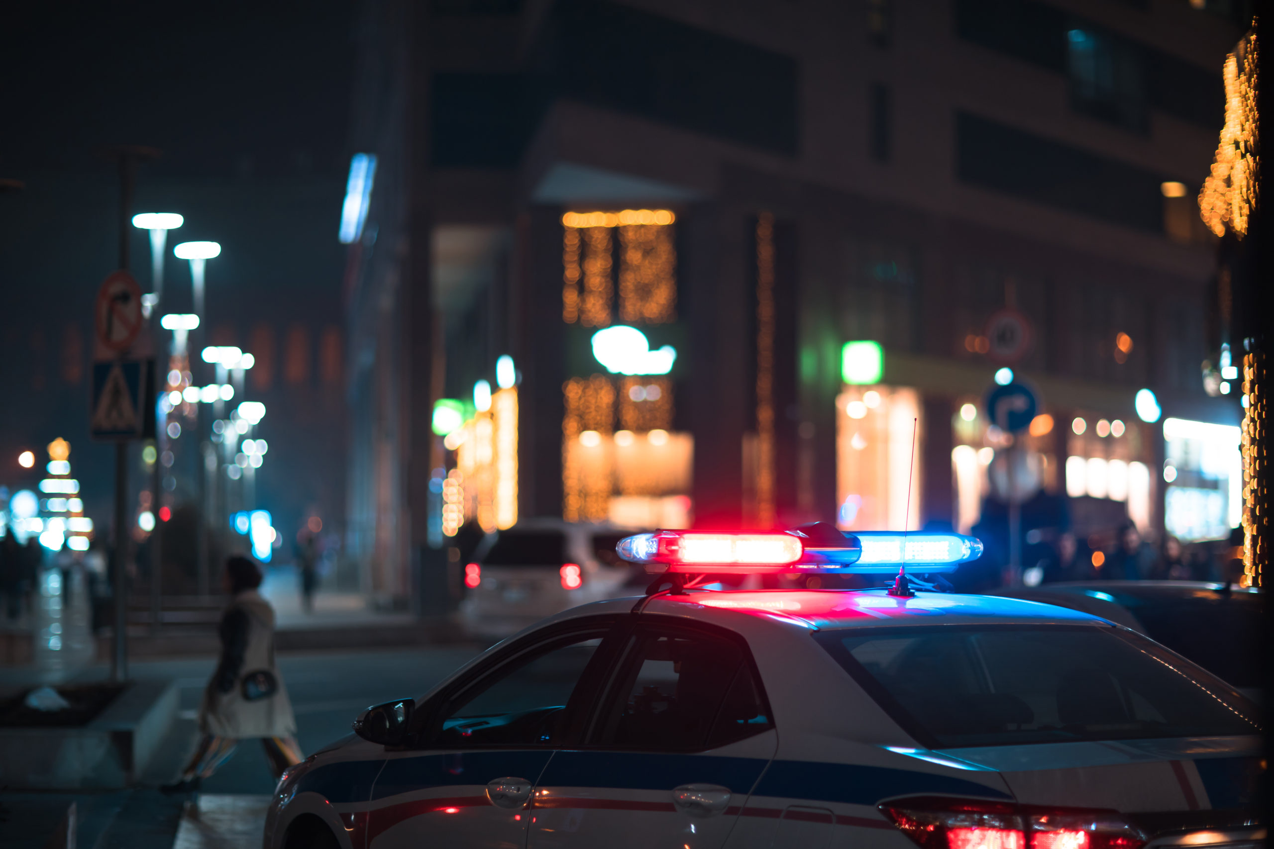 A police car with flashing red and blue lights is parked on a city street at night in Texas, casting an urgent glow. The brightly lit buildings and storefronts create an urban atmosphere. To the left, a person walks on the sidewalk, possibly pondering what to know about recent robbery charges.