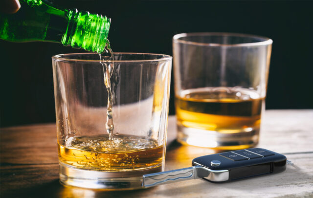 A green bottle pours liquid into a glass containing whiskey on a wooden table. Nearby, another glass with whiskey and a car key fob lies ominously, hinting at the serious DWI consequences in Texas, especially for those facing a second offense DWI.