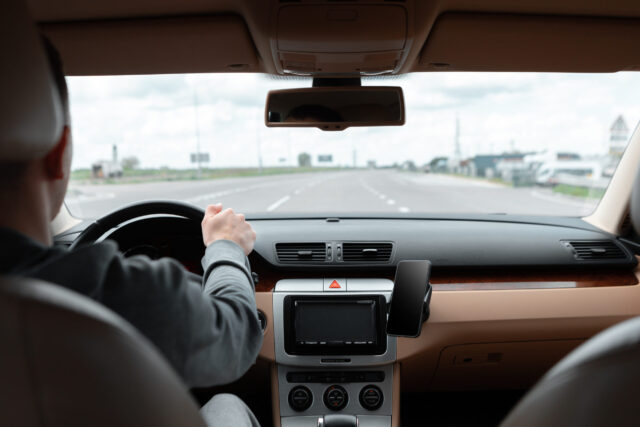 A person driving a car on a wide, empty Texas road. The view is from the backseat, showing the dashboard and steering wheel. The sky is cloudy, and the road stretches ahead with no vehicles in sight—an ideal scene for those who recently obtained their hardship license.