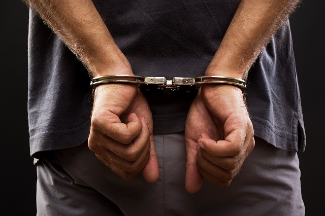 A person wearing a dark shirt and light pants is shown from the back, their hands cuffed with metal handcuffs. The dim background accentuates their restrained hands, hinting at possible criminal convictions.