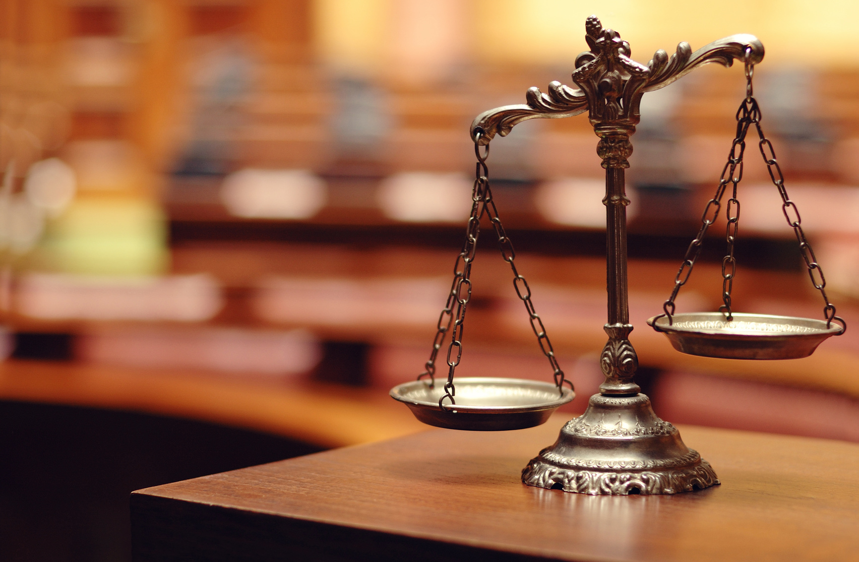 A close-up of a bronze balance scale, symbolizing justice, sits on a wooden surface in a TX courtroom. The blurred background with rows of seats and desks emphasizes the formal legal atmosphere, ready to address issues like contempt of court.