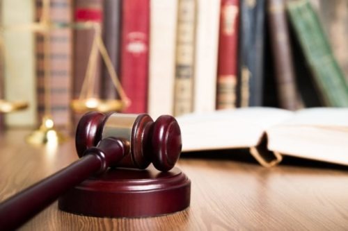 A judge's gavel rests on a wooden table, with an open book and a brass balance scale in the background. Books are lined on a shelf, indicating a legal or judicial setting. The focus is on the gavel in the foreground.
