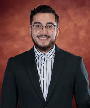 Demetris Guzmán, with short dark hair and glasses, is wearing a dark suit and a striped shirt, smiling against a textured reddish-brown background.