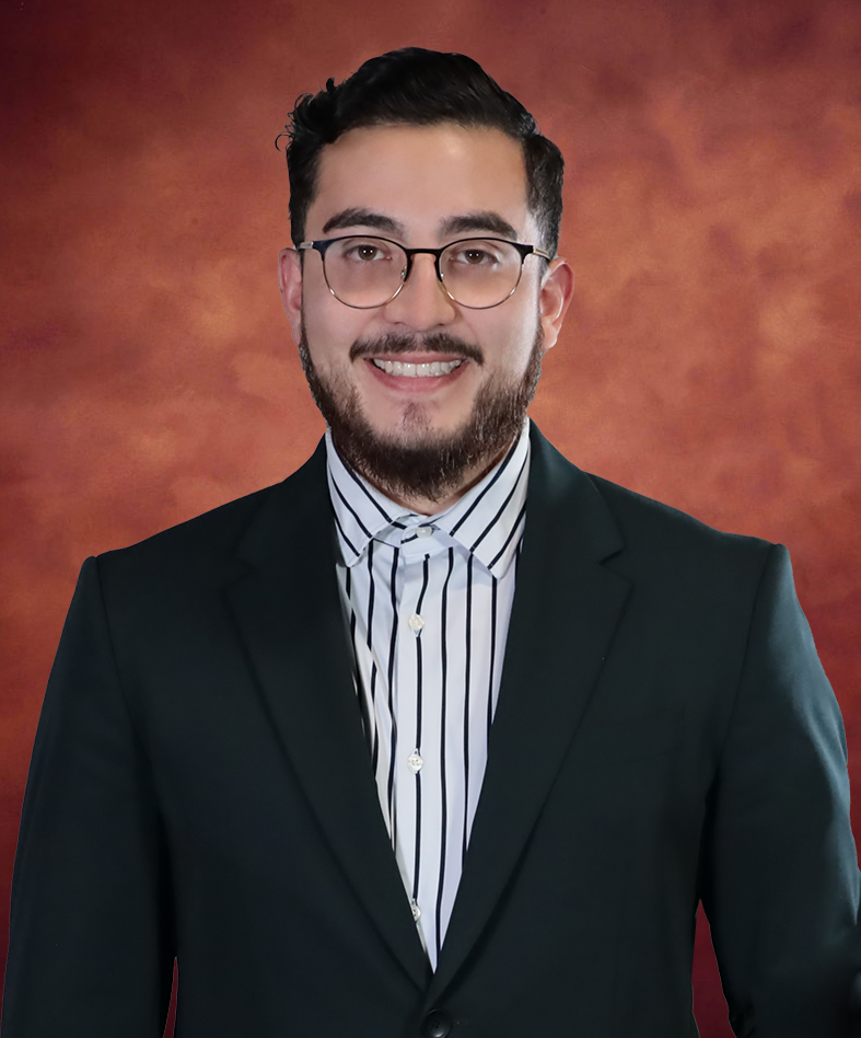 Demetris Guzmán, with short dark hair and glasses, is wearing a dark suit and a striped shirt, smiling against a textured reddish-brown background.