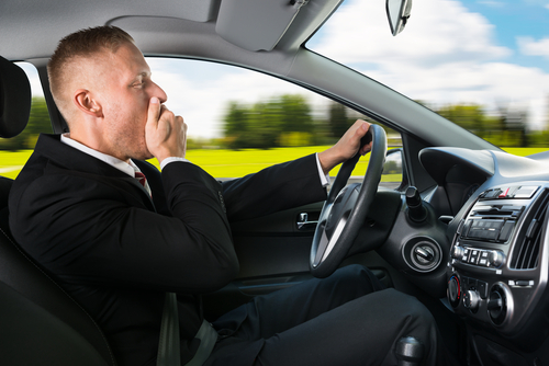 A man in a suit is driving through the vast Texas landscape, yawning as he covers his mouth—a subtle hint of driver negligence. The blurred green fields and blue sky whip by, while the car's interior, complete with a steering wheel and dashboard, adds to the sense of motion.
