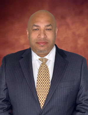 Faustino Morales, a bald man in a dark pinstripe suit and patterned yellow tie, stands confidently against a reddish-brown background. He wears a slight smile and sports a neatly trimmed goatee.