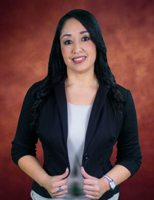 Gabby Martinez, a woman with long dark hair, smiles at the camera. She is wearing a black blazer over a light-colored top, her hands resting gracefully on her jacket. The background envelops her in a warm, gradient shade of brown.