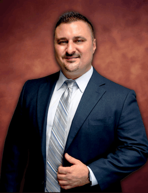 Marco Gonzalez, a man in a dark suit with a light striped tie, stands against a warm, reddish-brown background. He has a slight smile, short dark hair, and a goatee. With his right hand holding his jacket near the waist, he exudes quiet confidence.