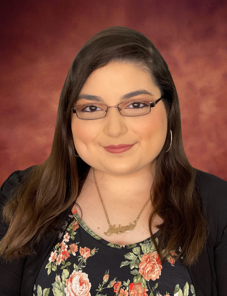 Portrait of Melanie Vasquez, with long brown hair and glasses, smiling softly. She wears a dark floral top paired with a necklace and a black cardigan against a warm, reddish-brown background.