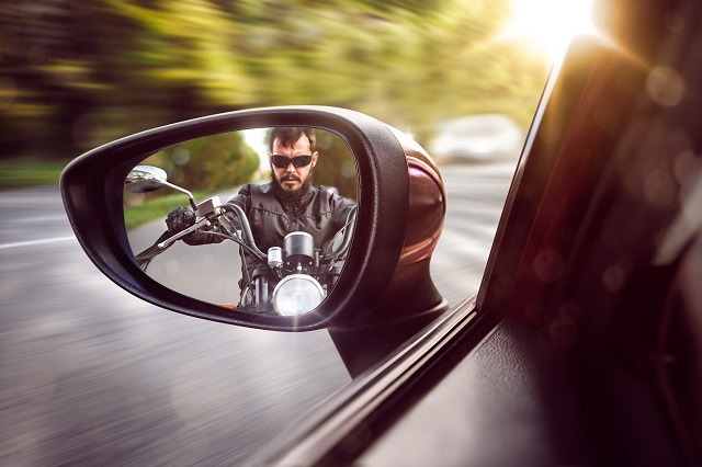 A man in a leather jacket and sunglasses rides a motorcycle, reflected in the side mirror of a moving car. Sunlight and blurred greenery fill the background, capturing his focus and speed—a reminder to know the steps to take for accident recovery should the unexpected happen.