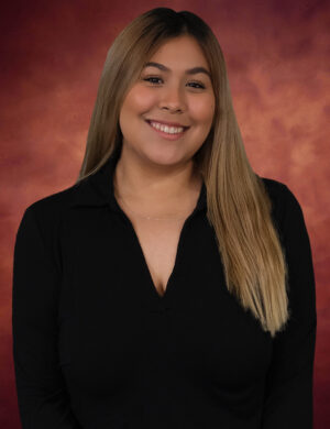 Perla Adilene Vazquez, with long, light brown hair, is smiling radiantly against a warm, blurred background. She's adorned in a black top and a delicate necklace.
