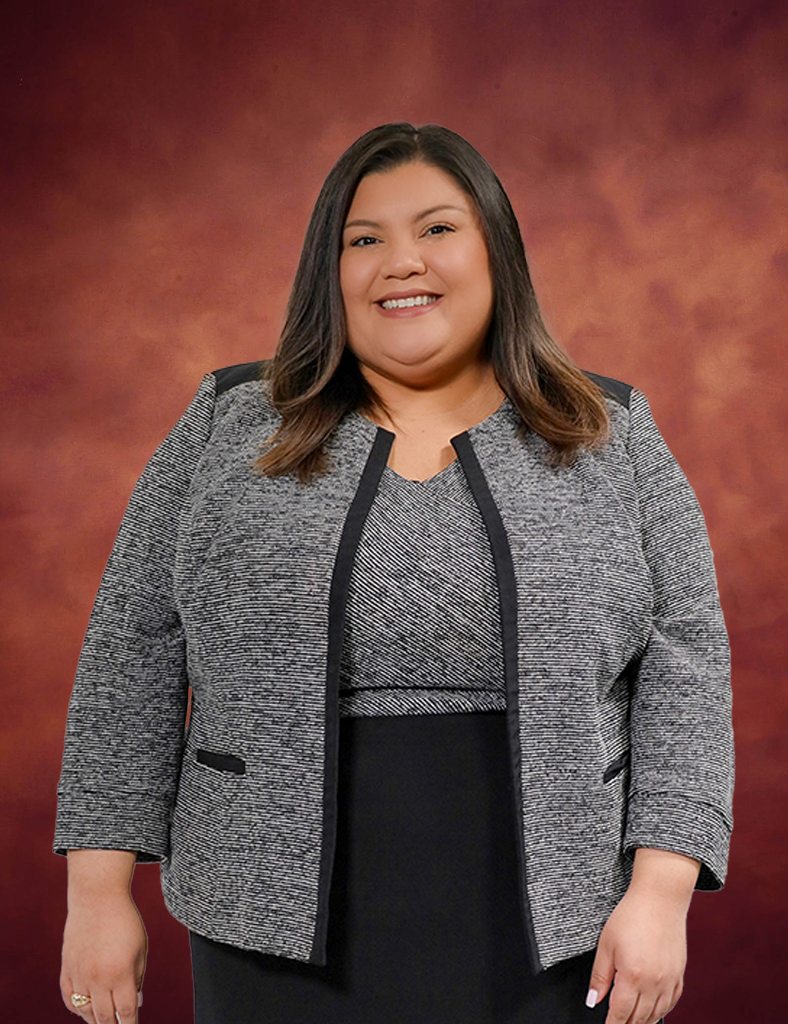 Marisa Ayala, with her long brown hair, smiles while wearing a gray blazer over a matching top and black skirt. She stands gracefully against a softly lit brown background.
