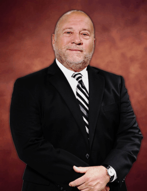 Ricardo N. Gonzalez, wearing a formal black suit and striped tie, stands against a warm, brownish-red background. His hands are clasped together as he looks directly at the camera with a neutral expression.