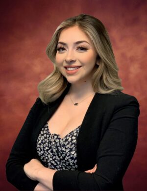 Stacey Zermeno stands with a warm smile, her long, wavy hair cascading over a dark suit jacket and patterned top. Her arms are confidently crossed against the backdrop of a rich, reddish-brown gradient.