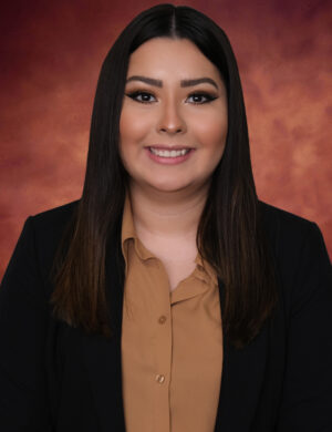 Daysi Umanzor, with her long, dark hair, smiles at the camera. She wears a black blazer over a tan shirt, perfectly complementing the warm, blurred blend of orange and red tones in the background.