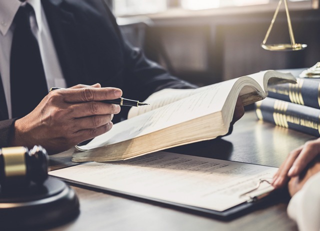 A person in a suit holds a pen and gestures towards an open law book on a desk. Nearby, another person listens with a clipboard. A gavel and scales of justice are visible, suggesting a legal setting like a meeting or consultation.
