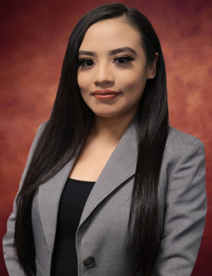 A woman with long black hair is smiling softly. She is wearing a gray blazer over a black top. The background is a warm, reddish-brown gradient.