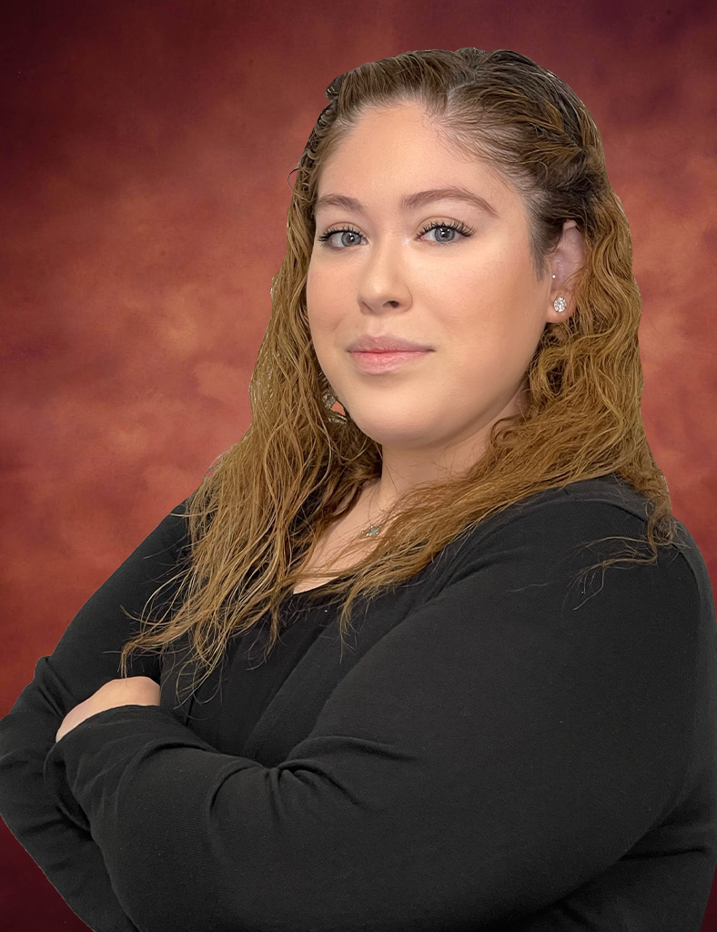 Yevette Vega, a woman with long, wavy hair, stands confidently with arms crossed in a black top. Set against a warm, reddish-brown background, she gazes directly at the camera.