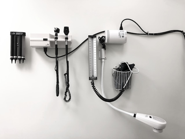Medical instruments mounted on a wall, including an otoscope, ophthalmoscope, blood pressure gauge, and thermometer. Cords are neatly organized. Against a plain white wall, these tools serve as reminders of Texas's commitment to combating healthcare fraud.