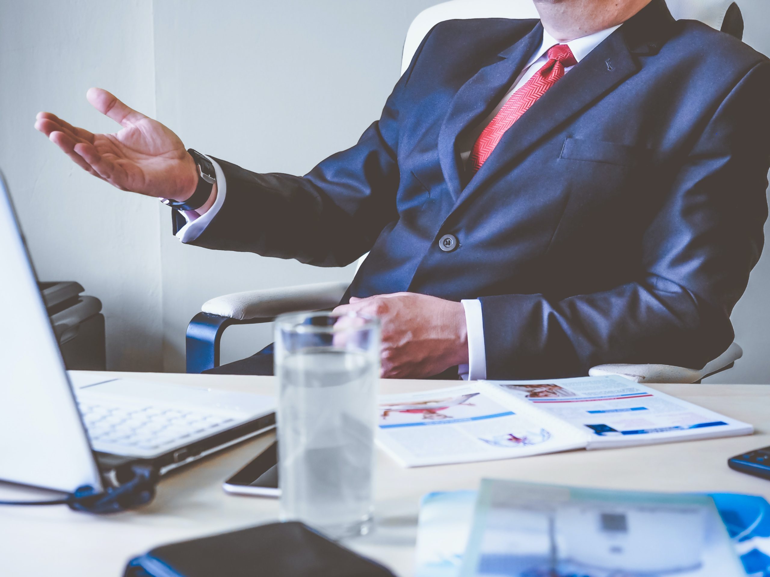 A criminal defense attorney in a business suit sits at a desk, gesturing with their left hand. On the desk are a laptop, smartphone, papers, and a glass of water. The focus is on the attorney's torso and hand as they strategize for their next big case.