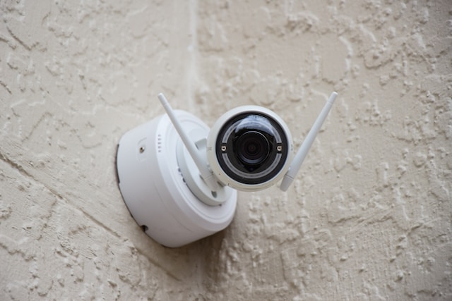 A white security camera with dual antennas is mounted on a textured, light-colored wall, vigilantly monitoring for any shoplifting incidents. The camera, reminiscent of the ones frequently seen in Texas stores to deter penalties, has its lens prominently visible as it faces forward.