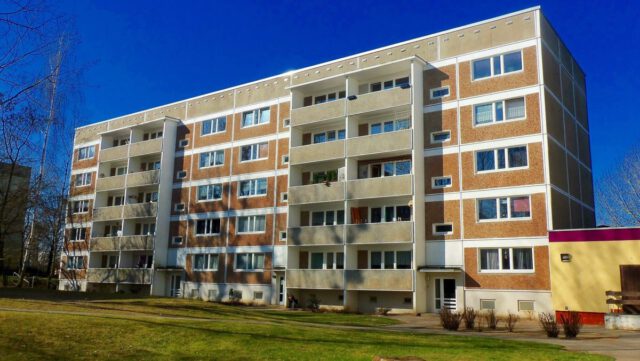 A five-story apartment building with beige and brown panels under a clear Texas sky. The facade features balconies, some with visible items, highlighting the need for apartment safety. In front, there's a grassy area and a few small trees. Shadows of branches are cast on the building.