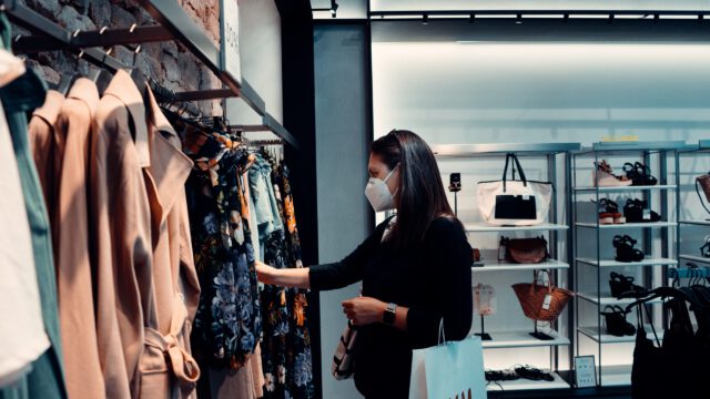 In a bustling Texas department store, a woman wearing a mask thoughtfully browses through a rack of patterned dresses. She clutches a white shopping bag while glancing at shelves lined with bags and accessories. It's important to stay vigilant about what to know regarding potential accidents while shopping.