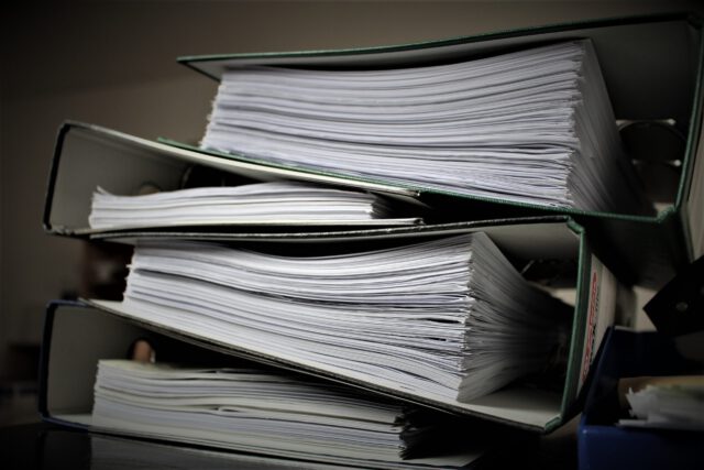 A close-up of a stack of thick binders filled with papers, with two binders laying horizontally on top of another. The binders are green and overflowing with white pages, suggesting a large volume of documents.