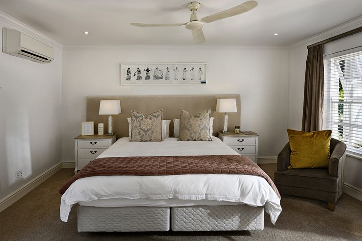 A cozy Airbnb bedroom features a large bed with plush pillows and a quilted headboard. Two bedside tables with lamps flank the bed, ensuring comfort. A ceiling fan and air conditioner provide ventilation, while a cushioned armchair with a yellow pillow invites relaxation by the window with blinds.