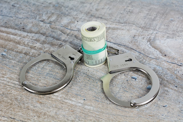 A pair of metal handcuffs lies open on a wooden surface, with a rolled stack of U.S. dollar bills secured by a green rubber band placed inside one cuff, hinting at the steep penalties associated with racketeering schemes.