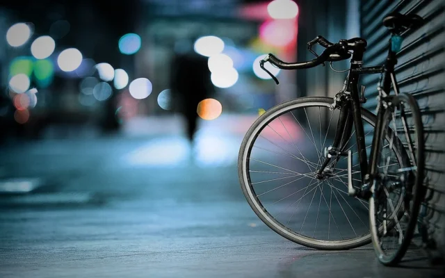 A bicycle with drop handlebars is propped against a metal fence on a dimly lit Texas street. Bokeh lights illuminate the background with a silhouette of a person walking in the distance. The scene conveys a serene, nighttime urban atmosphere, subtly hinting at what you should know about city life.