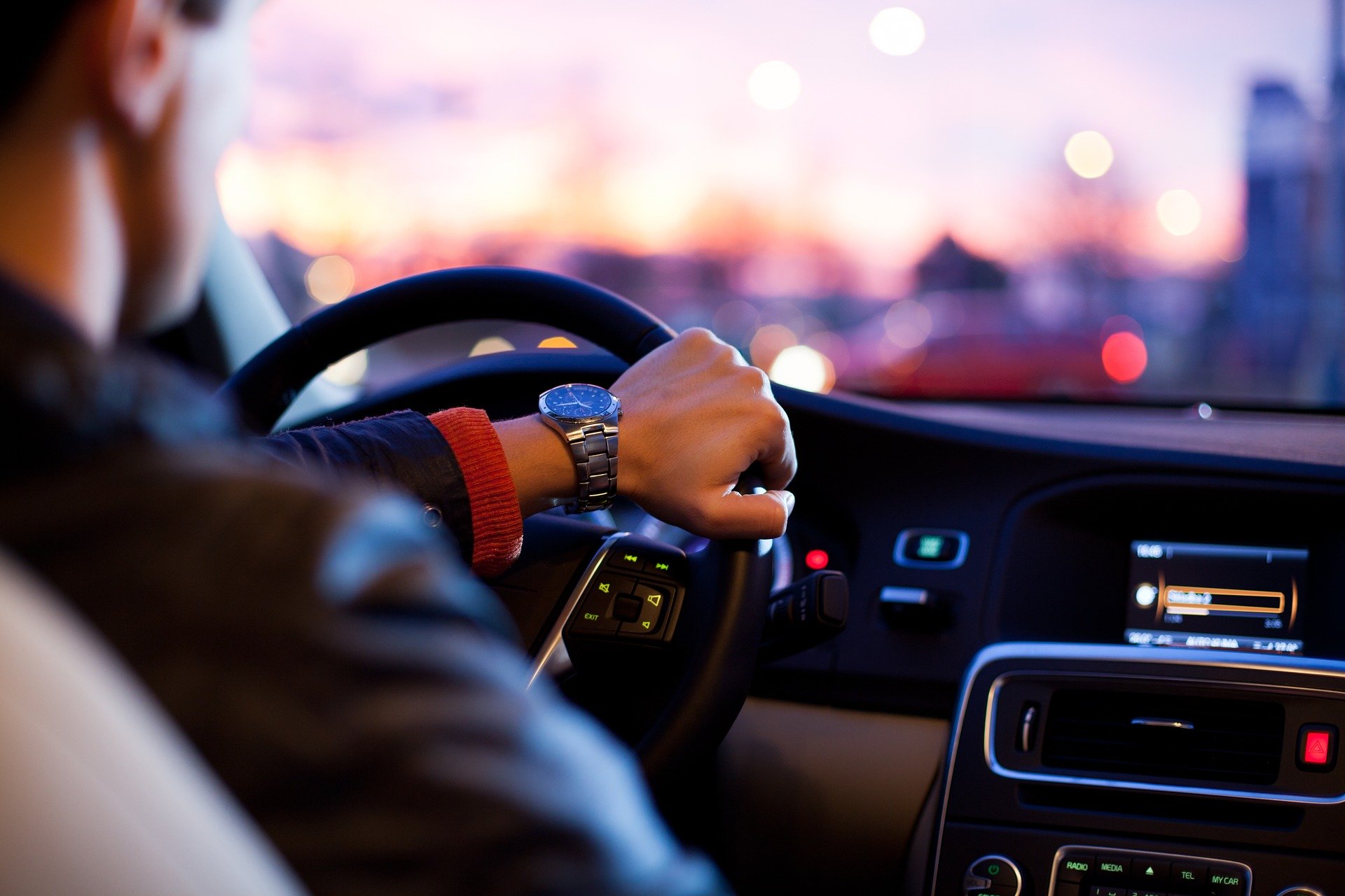 A person driving a car during sunset, with city lights visible through the windshield. The driver's left hand is on the steering wheel, wearing a watch, and the dashboard displays illuminated controls.