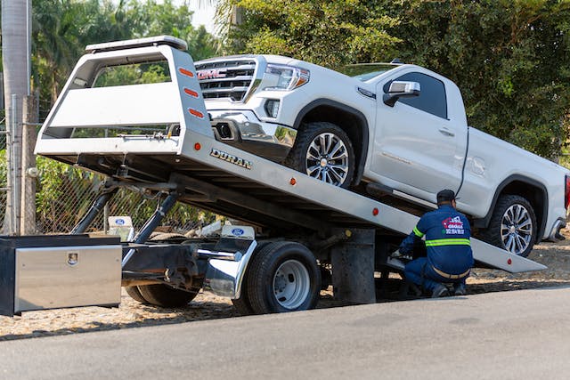 A white pickup truck is being impounded onto a tow truck's flatbed. A worker in a blue uniform kneels beside the tow truck, adjusting equipment. The background includes trees and a fence.