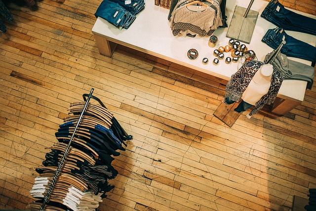 From above, a clothing store with a wooden floor reveals folded clothes on a white table and more on a circular rack. Small decorative items join the display next to a mannequin, setting an inviting scene where the staff discusses the consequences of shoplifting discreetly.